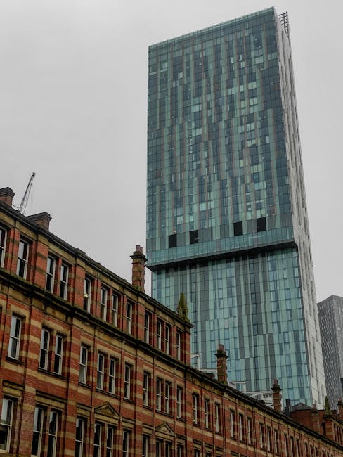 A large modern glass skyscraper with a reflective facade rising above traditional red-brick terraced houses in an urban area. The skyscraper features numerous vertical and horizontal lines, creating a