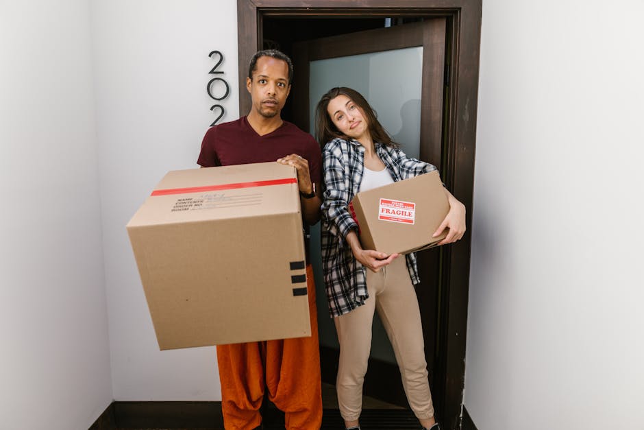 Two individuals are indoors, surrounded by large cardboard boxes and packing materials, preparing for a home relocation. The woman on the left is wearing casual attire, including a checked shirt and s